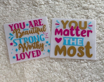 May include: Two white cards with inspirational messages in pink, blue, and gold lettering. The first card says "You are Beautiful Strong Worthy Loved." The second card says "You Matter The Most."