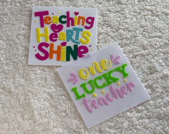 May include: Two colorful vinyl stickers with inspirational messages for teachers. The first sticker says "Teaching Hearts Shine" in pink, orange, yellow, and green letters. The second sticker says "One Lucky Teacher" in yellow, green, and pink letters.