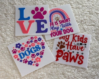 May include: Four vinyl stickers with dog-themed designs. The first sticker says "LOVE" in pink, blue, and purple letters. The second sticker says "Life is short, play with your dog" with a rainbow and paw print. The third sticker is a heart shape with paw prints and flowers surrounding the word "DOGS". The fourth sticker says "My kids have paws" with a brown paw print and red hearts.
