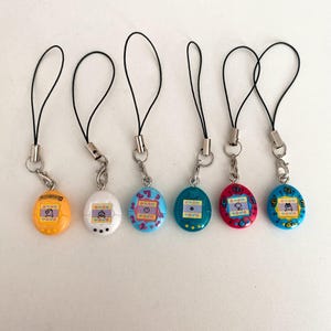 Puede incluir: Seis llaveros digitales de mascotas de colores con correas negras. Los llaveros son amarillos, blancos, azules, turquesas, rojos y azules. Cada llavero tiene una pequeña pantalla con un gráfico pixelado y botones.