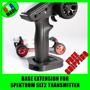 May include: Black remote control transmitter with a base extension. The transmitter has a black handle and a wheel on top. A small white and red toy car is in the background. The text "USA MADE" is in the upper left corner.