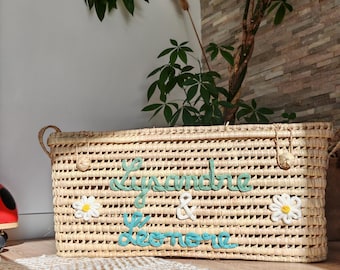 May include: Woven rectangular basket with handles, featuring the names "Lysandre & Léonie" in turquoise and green yarn. The basket is adorned with white daisy accents and sits on a textured rug, with a plant in the background.