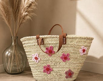 May include: Woven straw tote bag with brown leather handles and decorative pink crochet flowers. The bag is a light beige colour and has a wide opening. The bag is approximately 46 cm wide and 30 cm tall.