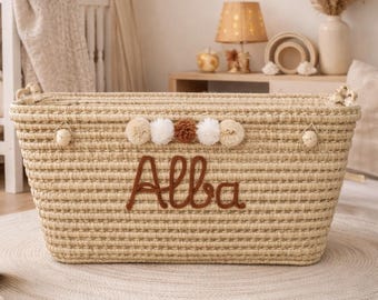 May include: Woven rectangular storage basket with the name "Alba" embroidered in brown thread. The basket is decorated with beige, white, and brown pom-poms. The basket is sitting on a round, beige rug.