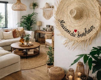 May include: A bright living room featuring a woven lampshade, a white sofa, and a wooden coffee table. A large straw hat with the text "Maison du bonheur" is mounted on the wall. The room is decorated with plants, woven baskets, and candles.