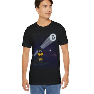 May include: A black t-shirt with a graphic of a cartoon Batman character standing in front of a shining Bitcoin symbol. The background is a dark blue night sky with stars.