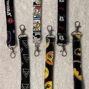 May include: A collection of six keychains with various designs. The keychains feature patterns including Spider-Man, Mickey Mouse, Donald Duck, emojis, and Batman. Each keychain has a silver-colored clasp.