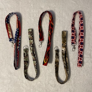 May include: Six different patterned lanyards with metal clasps. The lanyards feature a red, white, and blue American flag pattern, a camouflage pattern, a red and black flame pattern, a red and blue star pattern, and two different camouflage patterns.