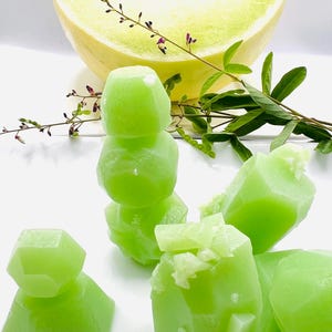 May include: A halved honeydew melon with visible seeds, alongside several bright green, geometric-shaped melon pieces. The arrangement includes a stack of melon cubes and scattered pieces, with sprigs of greenery and tiny purple flowers.