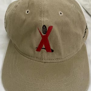 May include: A tan baseball cap with a red and black logo of a stick figure with a tennis racket. The logo says "X" in red.