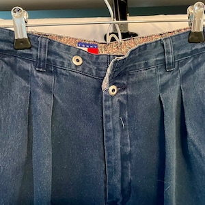 May include: Dark blue denim pants with pleats and a button closure. The pants have a patterned interior waistband and are hanging from a metal hanger. The brand name "TOMMY" is visible on a red tag.