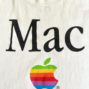 May include: A white t-shirt featuring the black text Mac printed in a serif font. Below the text is a graphic of an apple silhouette filled with horizontal stripes in green, yellow, orange, red, purple, and blue, with a small registered trademark symbol.