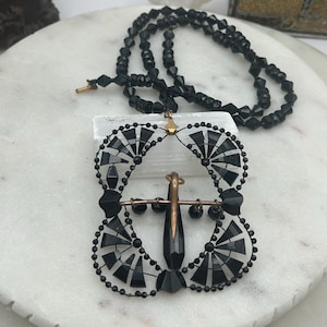 May include: A black beaded necklace with a pendant featuring a central cross design. The pendant is adorned with black geometric shapes and small black beads. The necklace is displayed on a white surface.