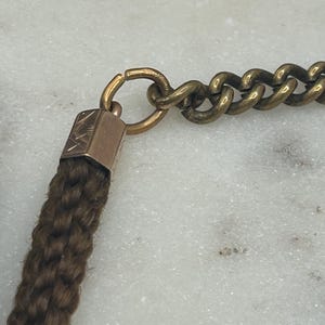 May include: Close-up of a vintage necklace featuring a woven brown braid, connected to a gold-toned metal chain. The chain is composed of interlocking links. The braid is attached to a small, rectangular gold-toned metal piece.