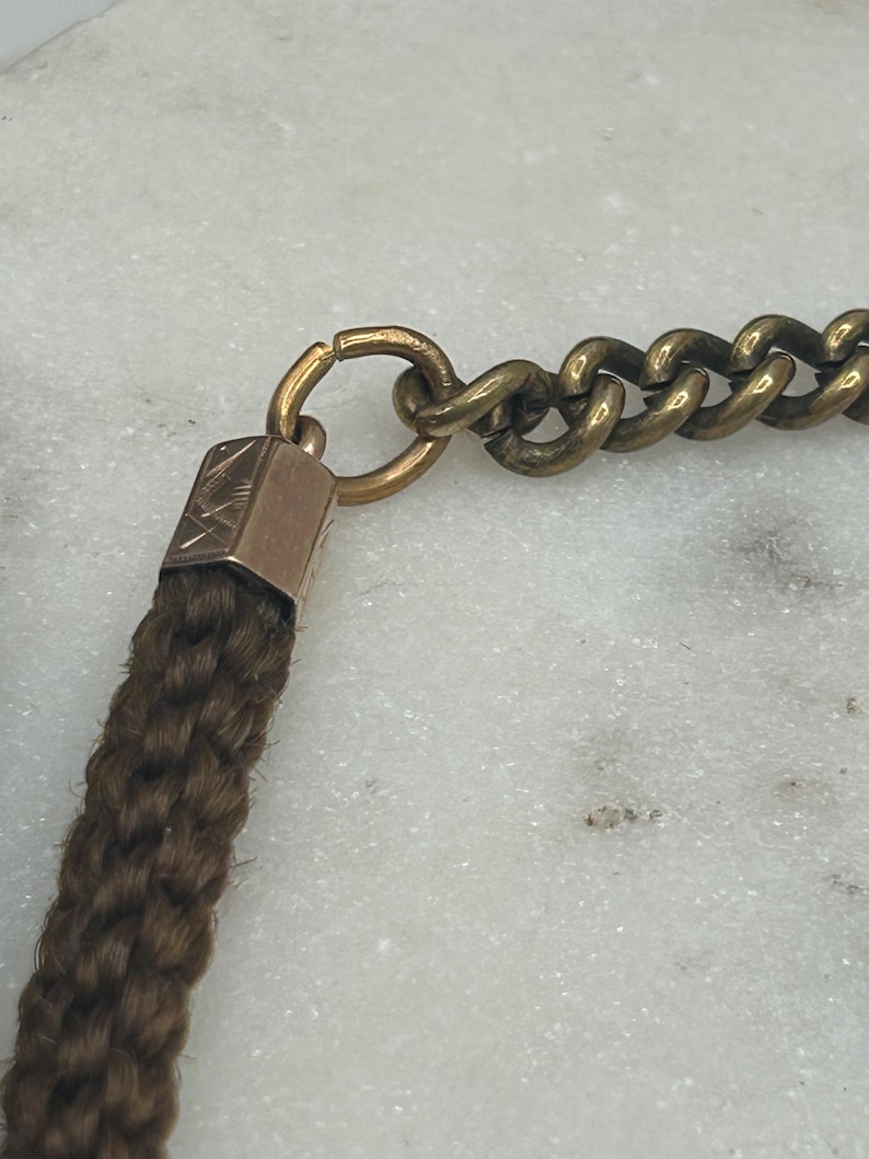May include: Close-up of a vintage necklace featuring a woven brown braid, connected to a gold-toned metal chain. The chain is composed of interlocking links. The braid is attached to a small, rectangular gold-toned metal piece.