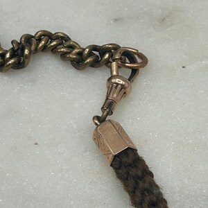 May include: Close-up of an antique watch chain with a gold-toned metal chain and clasp. The chain connects to a dark brown woven tassel. The background is a light-colored, textured surface.