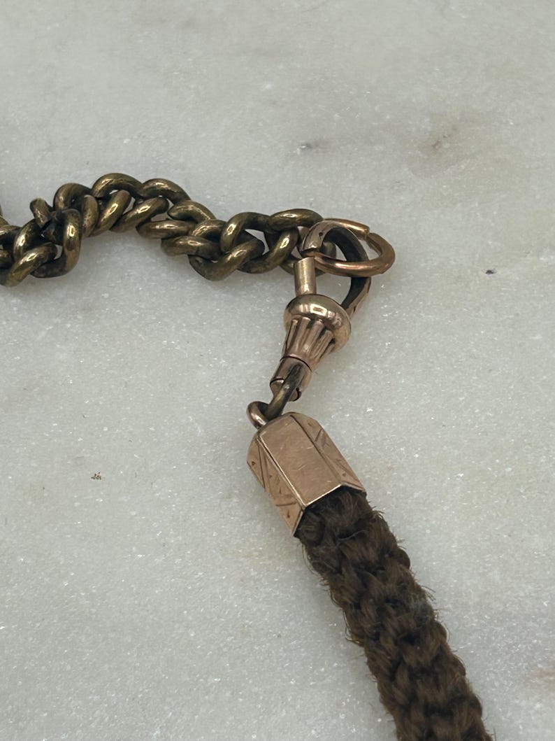 May include: Close-up of an antique watch chain with a gold-toned metal chain and clasp. The chain connects to a dark brown woven tassel. The background is a light-colored, textured surface.
