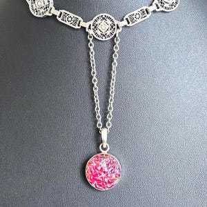 May include: Silver-toned necklace with a filigree design. The necklace features a series of geometric shapes and a pendant with a round, pink, and sparkly center. The necklace is displayed against a dark background.