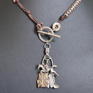 Può includere: Una collana con un cordino in pelle marrone decorato con perline metalliche e una chiusura a T argentata. Dalla chiusura pende un ciondolo argentato che raffigura un cavallo con cavaliere che salta sopra un paio di stivali da cowboy.