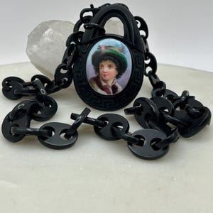 Puede incluir: Collar vintage negro con un colgante central que representa un retrato de una persona con sombrero. El colgante está enmarcado en negro y conectado a una cadena. El collar se exhibe sobre una superficie blanca.