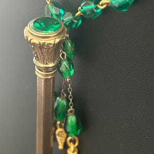 Op de afbeelding: Een vintage potloodhanger ketting. De goudkleurige potlood heeft een grote groene edelsteen aan de bovenkant. De ketting heeft groene facetkralen en bungelende groene kralen accenten. Het potlood is ongeveer 10 cm lang.