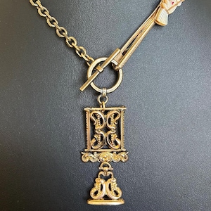 Puede incluir: Collar dorado con un colgante decorativo. El colgante presenta un marco rectangular adornado con detalles en espiral y un elemento inferior en forma de campana. Una cadena dorada y un cierre de barra con un pequeño acento blanco y rojo completan el diseño.