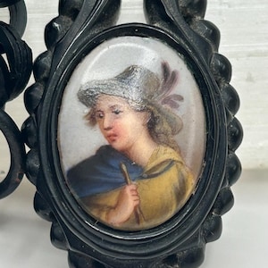 May include: A black oval pendant with a miniature portrait of a person wearing a hat and yellow clothing. The portrait is set within a decorative frame and attached to a black chain.