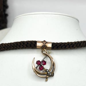 May include: A vintage gold-tone pendant necklace featuring a crescent moon design with three red gemstones and small pearls. The pendant hangs from a dark brown braided cord. The necklace is displayed on a white mannequin bust.