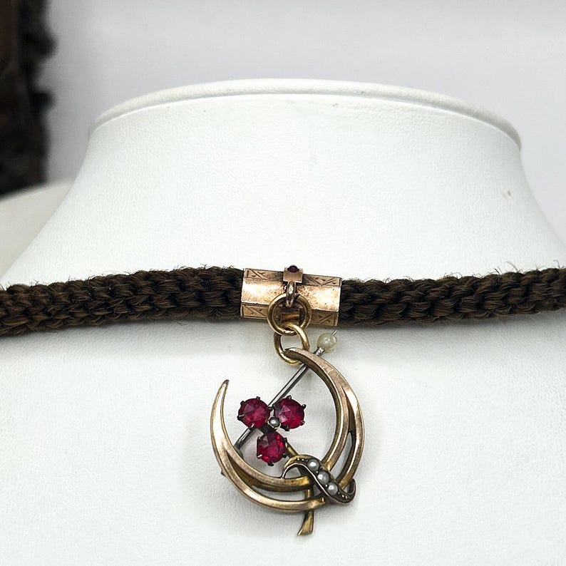 May include: A vintage gold-tone pendant necklace featuring a crescent moon design with three red gemstones and small pearls. The pendant hangs from a dark brown braided cord. The necklace is displayed on a white mannequin bust.