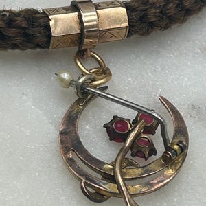 May include: Close-up of an antique necklace featuring a braided brown cord with gold-toned metal accents. A crescent-shaped pendant with three red gemstones and a small pearl hangs from the cord, showcasing a vintage aesthetic.