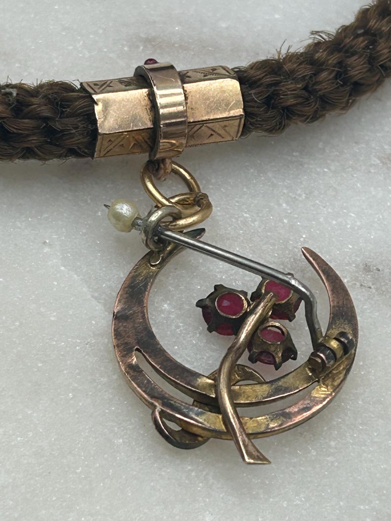 May include: Close-up of an antique necklace featuring a braided brown cord with gold-toned metal accents. A crescent-shaped pendant with three red gemstones and a small pearl hangs from the cord, showcasing a vintage aesthetic.