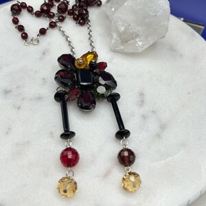 May include: A silver chain necklace with a black and red crystal flower pendant. The pendant has two black dangling strands with red and yellow crystal beads.