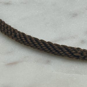 May include: Close-up of a section of brown rope. The rope is tightly woven, with a textured appearance. The rope is set against a white and gray marble background. The rope is a dark brown color.