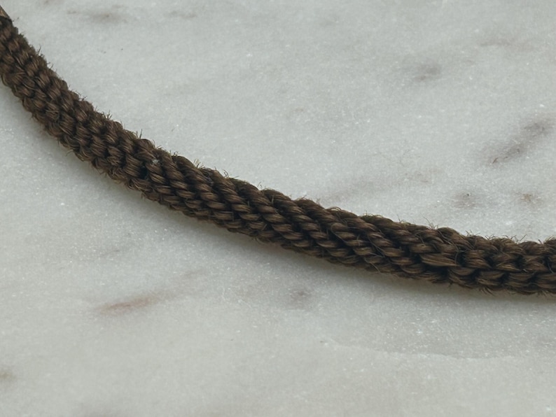 May include: Close-up of a section of brown rope. The rope is tightly woven, with a textured appearance. The rope is set against a white and gray marble background. The rope is a dark brown color.