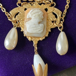 May include: Gold necklace with a cameo pendant depicting a woman's profile. The pendant is set in an ornate gold frame and has pearl-shaped drops. The necklace has a delicate chain and is set against a purple background.