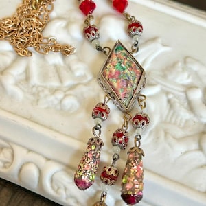 May include: Vintage-style necklace with a gold chain and ornate pendant. The pendant features a diamond-shaped, iridescent center framed in silver. Red and gold beads accent the chain and dangle elements, creating a glamorous, antique aesthetic.