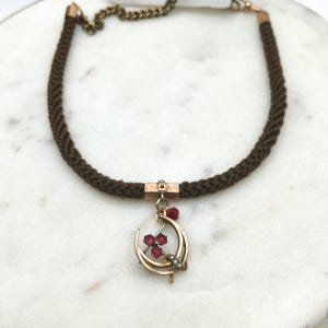 May include: A vintage necklace featuring a dark brown braided cord. The necklace has a gold-toned crescent moon pendant with three red gemstones and small pearls. A small gold-toned chain connects the necklace.