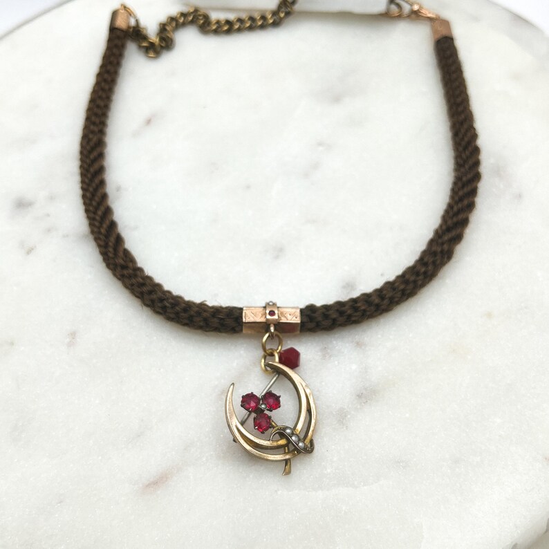 May include: A vintage necklace featuring a dark brown braided cord. The necklace has a gold-toned crescent moon pendant with three red gemstones and small pearls. A small gold-toned chain connects the necklace.