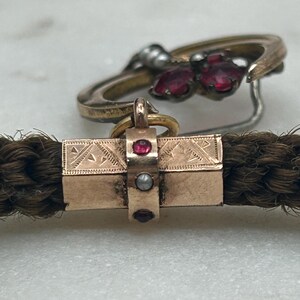 May include: Close-up of antique jewelry featuring a braided hair bracelet with a gold clasp adorned with red gemstones and a pearl. A gold ring with red gemstones is in the background. The jewelry is set against a marble surface.