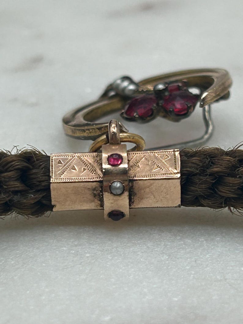 May include: Close-up of antique jewelry featuring a braided hair bracelet with a gold clasp adorned with red gemstones and a pearl. A gold ring with red gemstones is in the background. The jewelry is set against a marble surface.