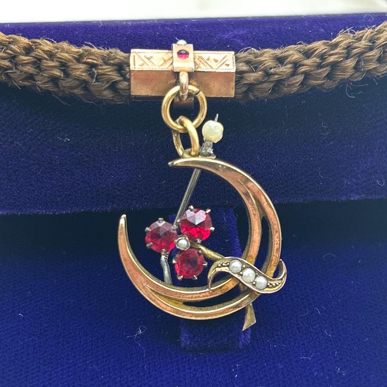 May include: A gold-toned brooch with a crescent moon design, featuring three red gemstones and small pearls. The brooch is suspended from a rectangular bar with etched details and a small red gemstone. The background is a deep blue velvet.
