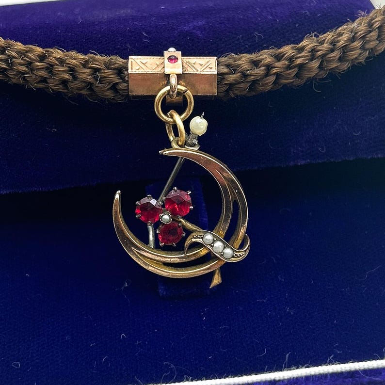 May include: A gold-toned pendant featuring a crescent moon design with three red gemstones and small pearls. The pendant is suspended from a rectangular bar with a small red gem and a braided brown cord.