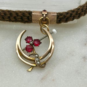 May include: A vintage gold-toned pendant with a crescent moon design, adorned with three red gemstones and small pearls. The pendant is attached to a brown braided cord. The pendant is likely an antique piece of jewelry.