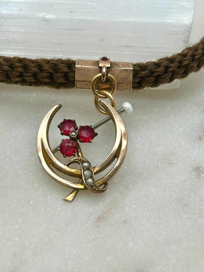 May include: A vintage gold-toned pendant with a crescent moon design, adorned with three red gemstones and small pearls. The pendant is attached to a brown braided cord. The pendant is likely an antique piece of jewelry.