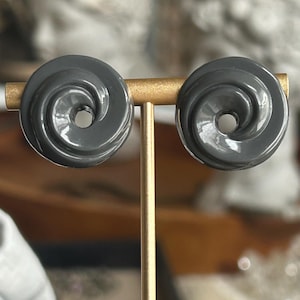 May include: A pair of gray swirl-design stud earrings. Each earring features a spiral pattern with a central hole. The earrings are displayed on a gold-colored stand, showcasing their unique design and shape.