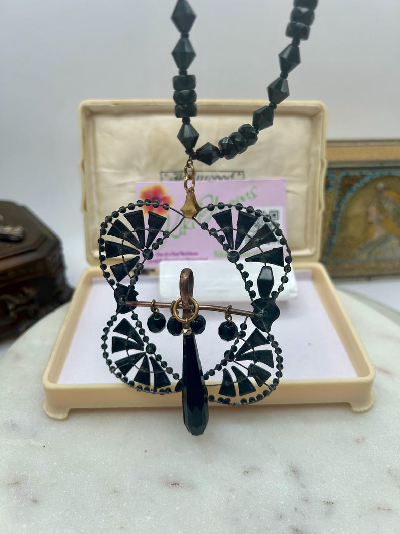 以下が含まれることがあります： 扇形のデザインのペンダントが付いた、ヴィンテージの黒いビーズネックレス。ペンダントには、涙型の黒い宝石と小さな黒いビーズが付いています。ネックレスはクリーム色の箱に入っています。