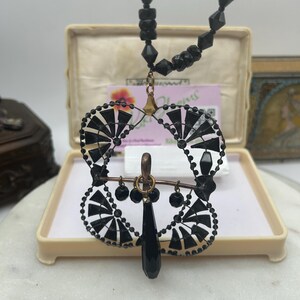以下が含まれることがあります： ディスプレイボックスに入った、ヴィンテージの黒いビーズネックレス。ペンダントは、黒と白のアクセント、涙型のビーズ、小さなぶら下がりビーズを備えた扇のようなデザインが特徴です。ネックレスは白い表面に展示されています。