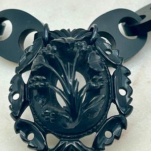 Può includere: Una collana con pendente nero intagliato in modo intricato. Il pendente presenta un design ovale centrale con motivi floreali e di foglie. La catena della collana è composta da maglie grandi e arrotondate. Il design generale è ornato e dettagliato.