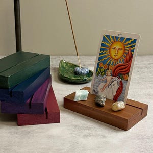 May include: A stack of four rectangular wooden card stands in green, blue, purple, and red. A single brown wooden stand holds a tarot card depicting a sun, a child, and a white horse. Three small raw crystals sit on the brown stand near the card.