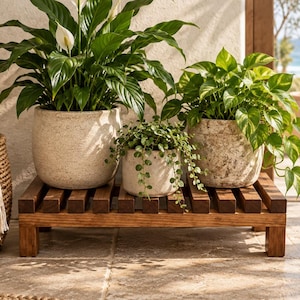 May include: A rectangular wooden plant stand with a slatted top surface holds three potted plants. The left pot is large and speckled, the center pot is small and light-colored, and the right pot is medium-sized with a textured, weathered finish.